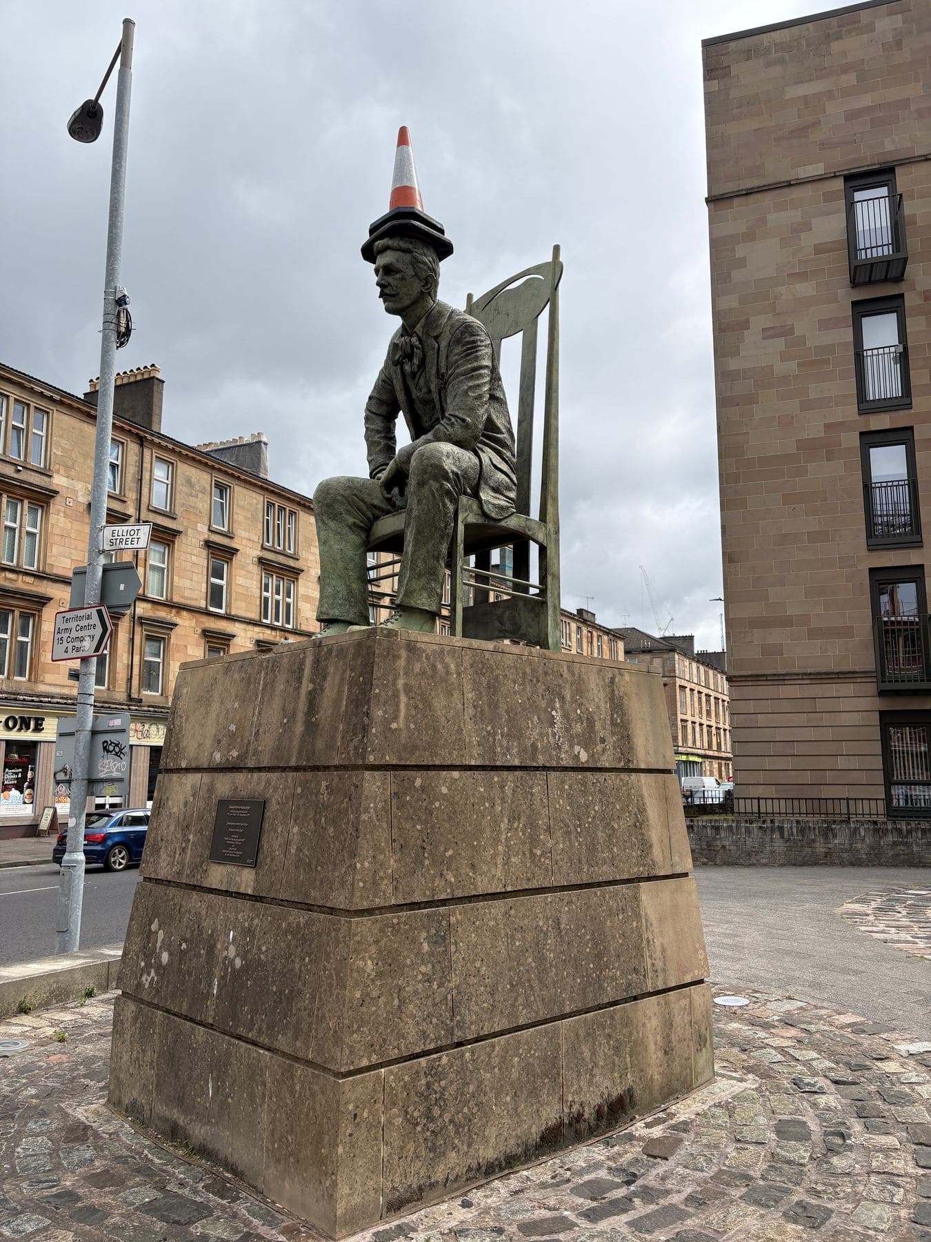 Statue of Charles Rennie Mackintosh seated on a chair, on a stone plinth
