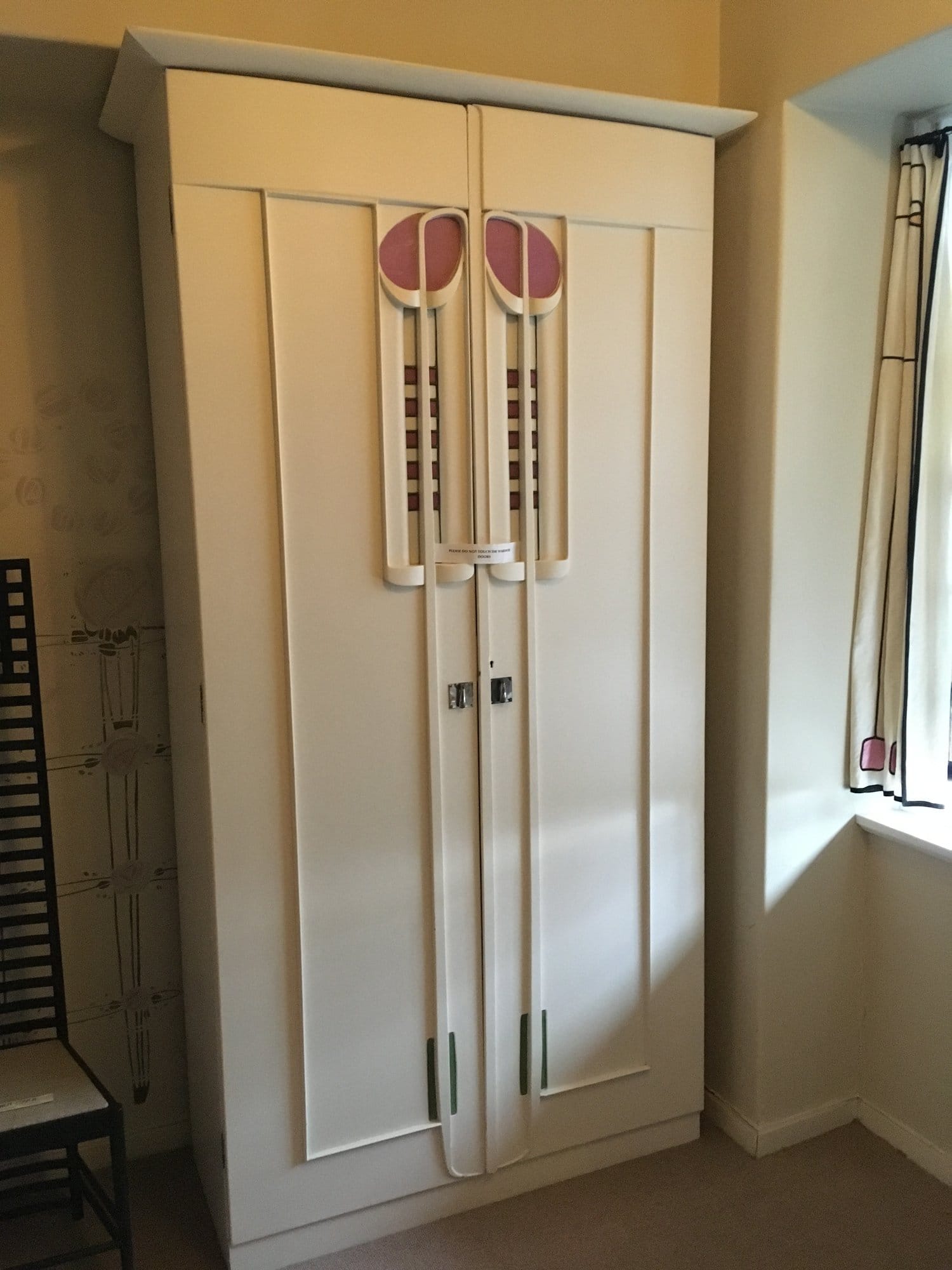 A white wardrobe with a rose shape and long stems on its doors, beside a tall ladder-back chair