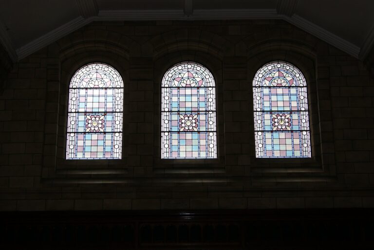 Row of three arched windows glazed in pale pink, blue and cream leaded quarries in a dim room