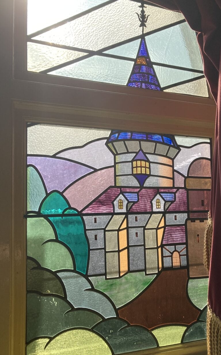 A coloured leaded window with a castle tower on a hill, with rounded green hills in the foreground.