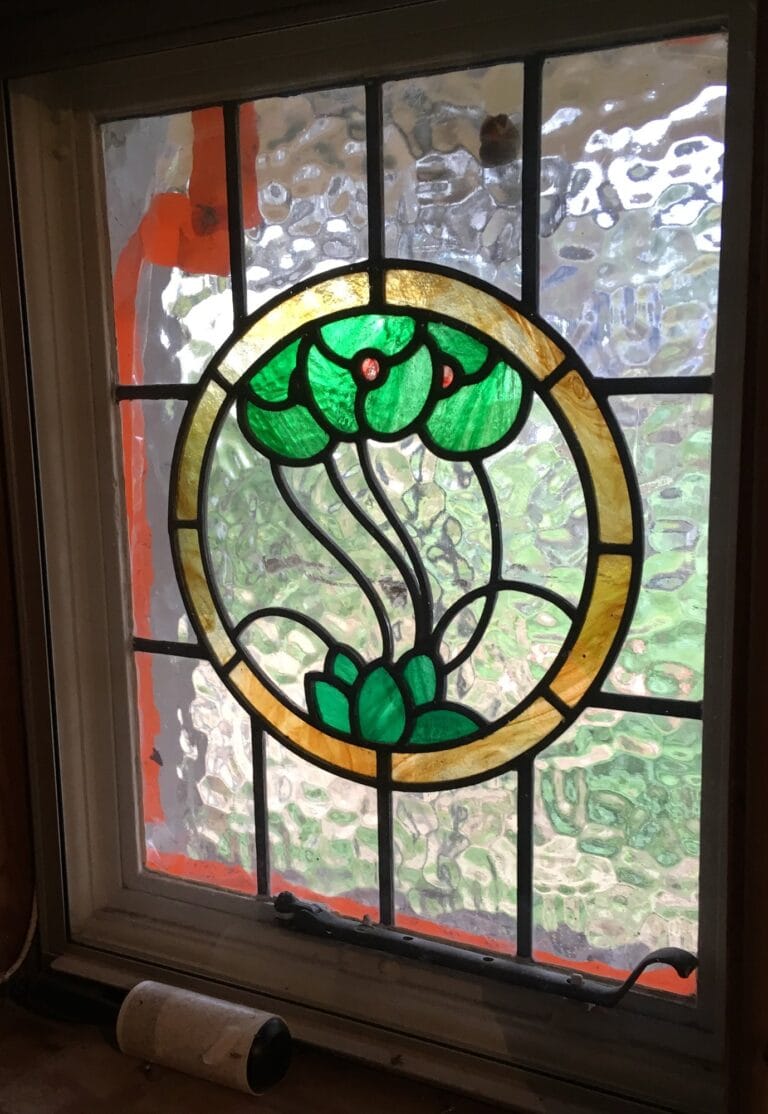 A round panel of green leaves and flowers in a yellow ring, set in textured glass in a wooden window.