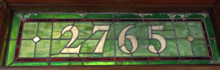 A leaded transom above a wooden door with the number 2765 in pale glass on a green background with a red border.