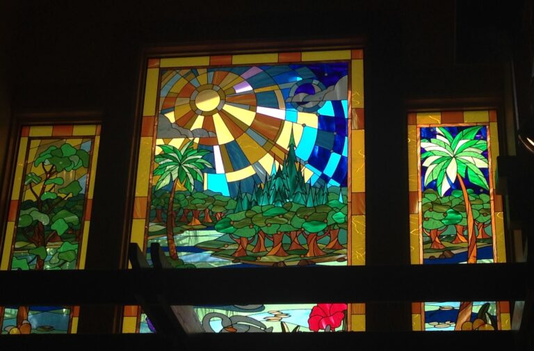 A large coloured window with a sunburst over palm trees and water, with a narrower panel of trees and plants on each side.