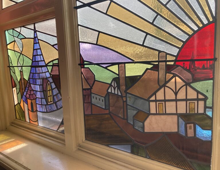 A coloured leaded window with a half-timbered house, a church spire and a red sun rising behind rolling hills, in a wide window with a central bar.